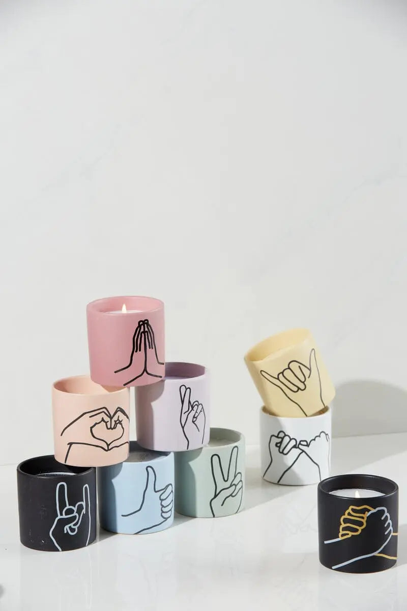 The Tobacco + Vanilla "Love Ya" - Impressions 5.75 oz Candle features a hand gesture illustration on its jar, displayed in a vibrant arrangement with other scented candles on a white surface.