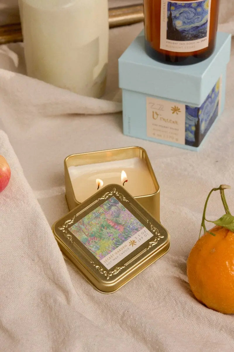 A Claude Monet Secret Garden - Impressionist 3.5 oz Tin Candle glows in a gold tin on beige fabric, beside citrus fruits and a blue Van Gogh-inspired candle—perfect for artful, scented gift sets.