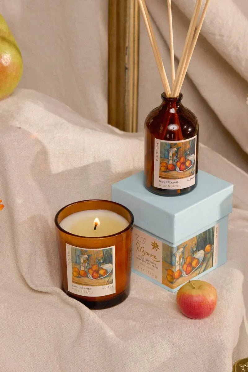 A Cezanne Wild Neroli Impressionist Diffuser (2.7 fl oz) in a brown glass bottle sits on a blue box with fruit-themed labels; nearby are an apple and beige fabric—perfect artistic gifts for any space.