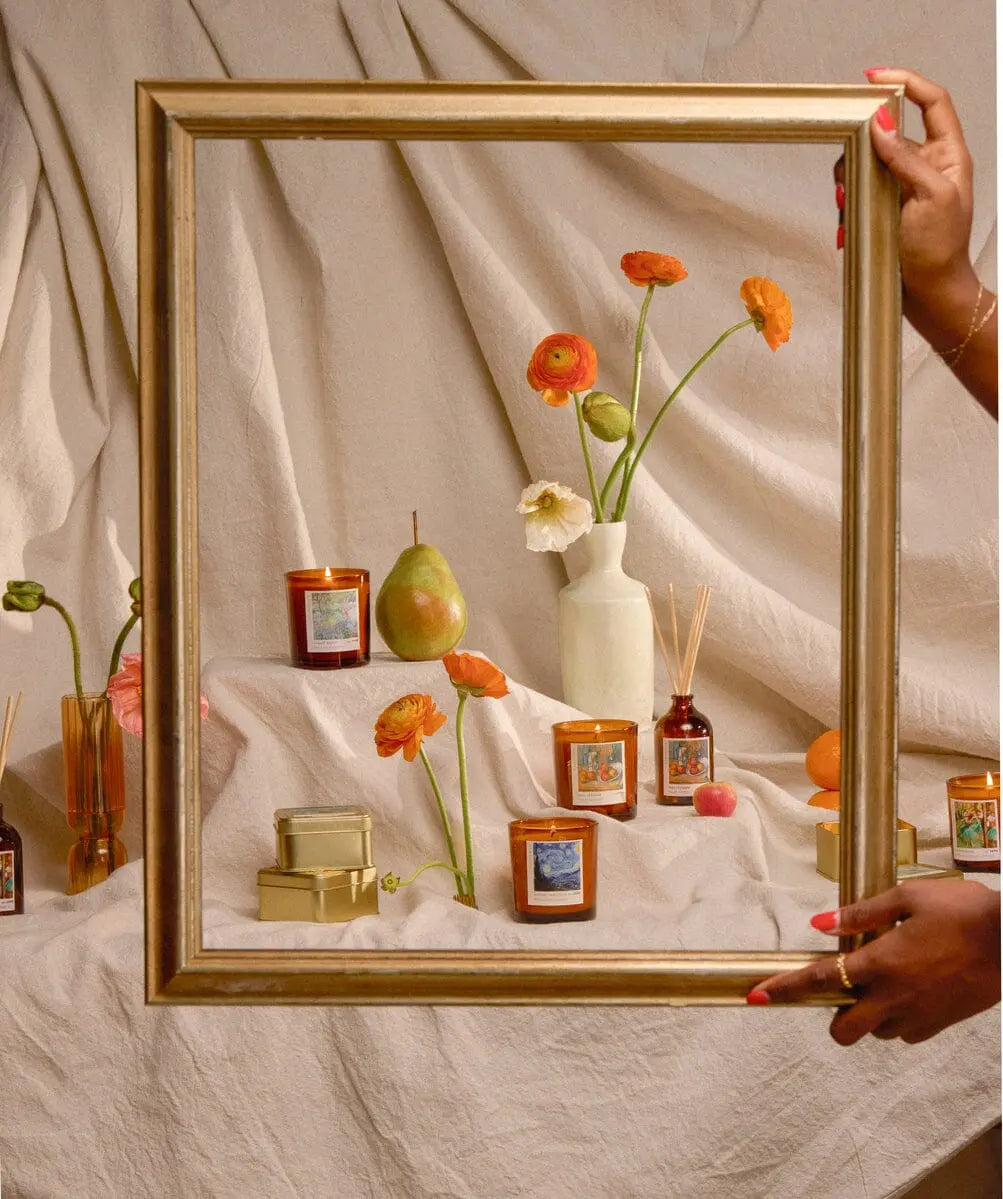 Hands hold a gold frame in front of Vincent Midnight Cedre - Impressionist Diffuser 2.7 fl oz, candles, flowers in a white vase, a pear, and small boxes arranged on draped beige fabric.