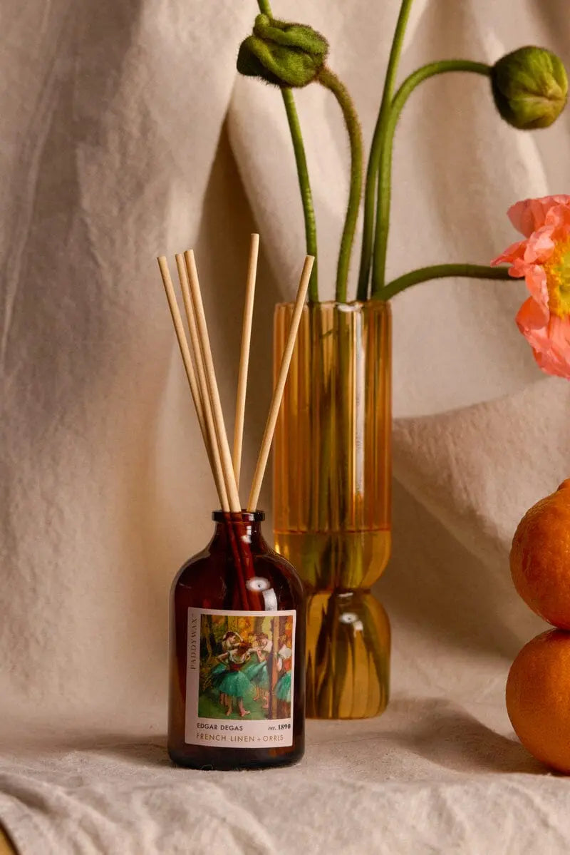 The Degas French Linen + Orris Impressionist Diffuser (2.7 fl oz) in a brown glass bottle with wooden reeds stands before an amber vase of flowers and two oranges, creating an inviting, art-inspired fragrance display on a fabric surface.