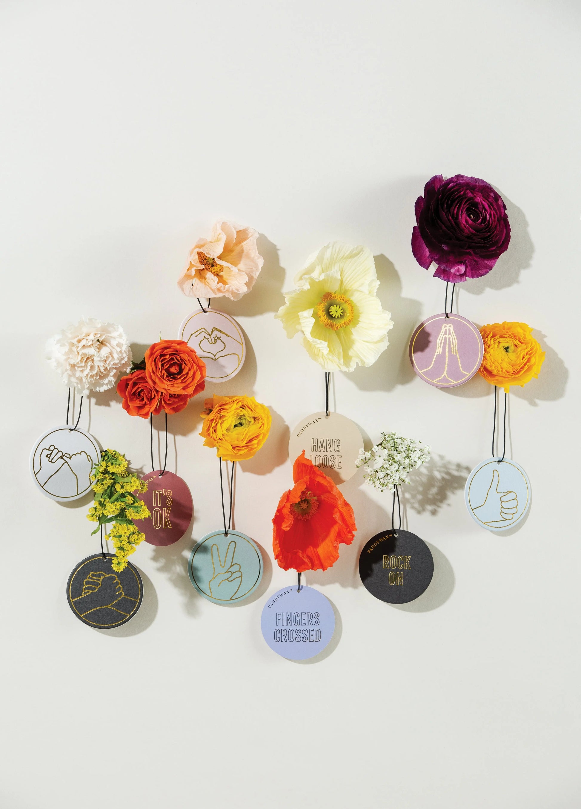 Circular embroidery hoops with hand gesture designs and phrases like "Pinky Promise" hang on a wall, each with colorful artificial flowers, creating a display as charming and unique as our Wild Fig + Cedar "Pinky Promise" car fragrance.
