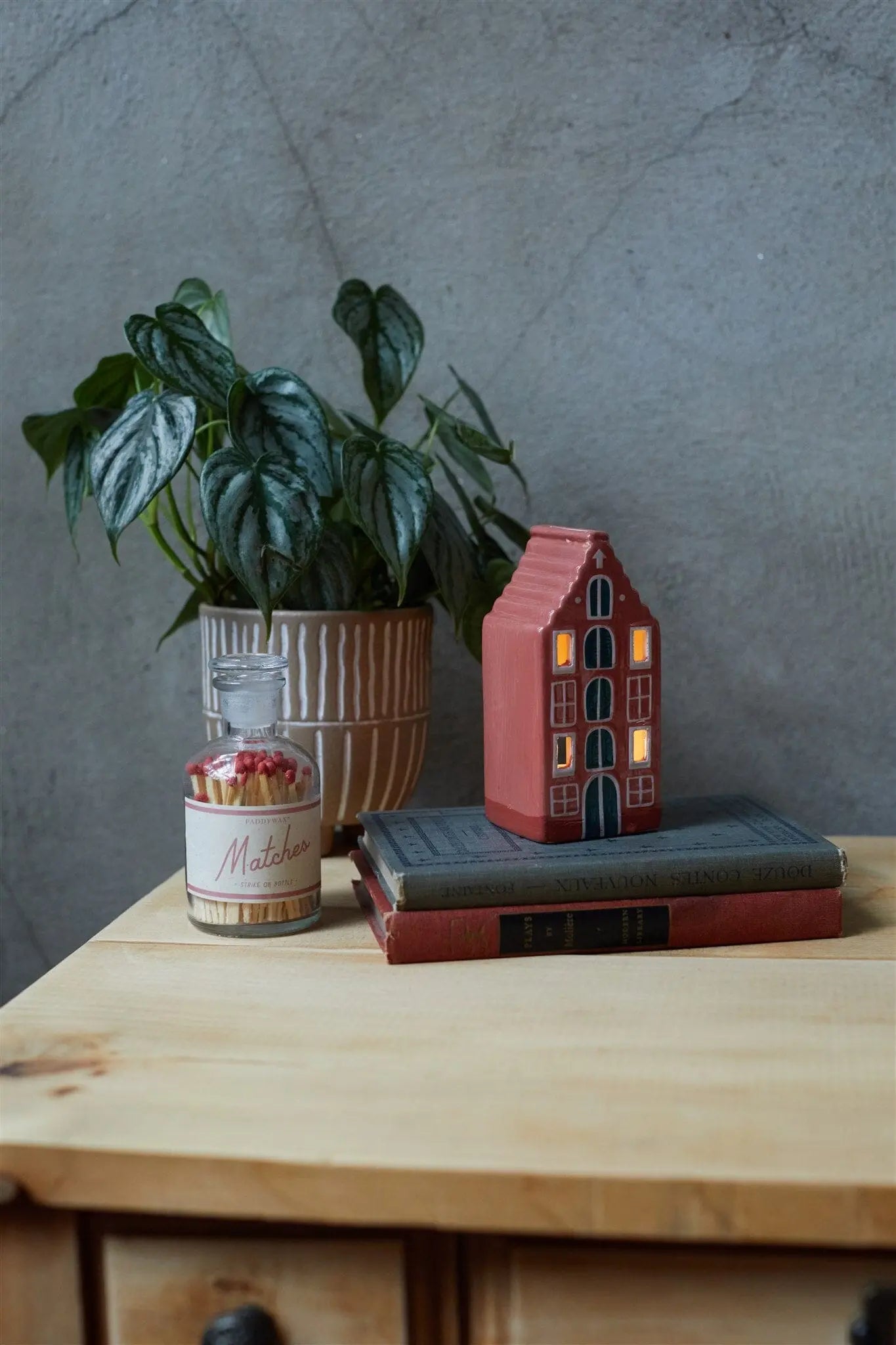 A potted plant, a jar labeled Matches, and the No. 02 The Amsterdam House – Incense & Tea Light Holder sit on two stacked books atop a wooden table against a gray wall.