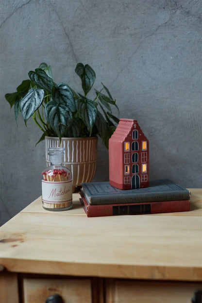 A potted plant, a jar labeled Matches, and the No. 02 The Amsterdam House – Incense & Tea Light Holder sit on two stacked books atop a wooden table against a gray wall.