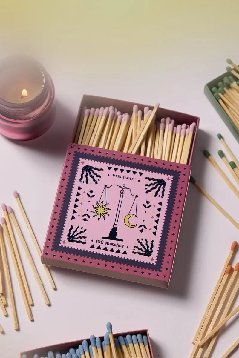The Balance - Pink Matches set, featuring pastel jumbo matches in open matchboxes with a scales illustration, is arranged alongside a pink candle on a white surface.