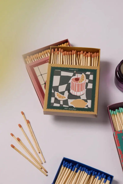 Three open boxes of Cocktail - Orange Matches with colorful jumbo matches are displayed on a white surface; one box shows a drink, lemon slices, and a patterned background, making it a stylish candle accessory.