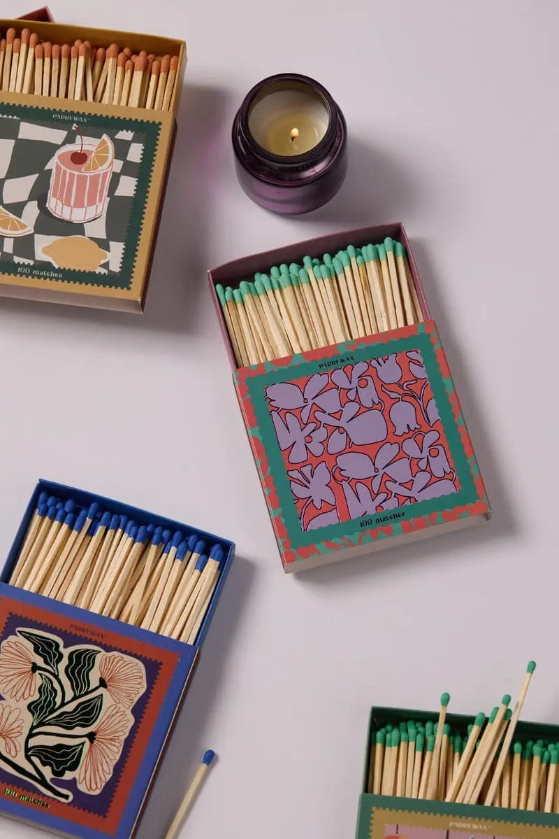 Four Abstract Floral - Teal Matchboxes filled with jumbo, vibrant-tipped matches are arranged around a lit purple candle—an eye-catching accessory for any light surface.