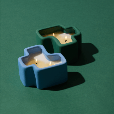 Two lit candles in geometric ceramic holders, one blue and one green, are placed on a green surface with distinct shadows.