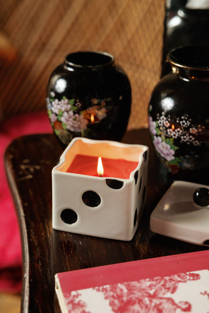 A lit Lava Stone - Lucky Dice 10 oz Candle in a white, die-shaped keepsake box with black spots sits on a wooden table next to its lid and two small black floral vases.