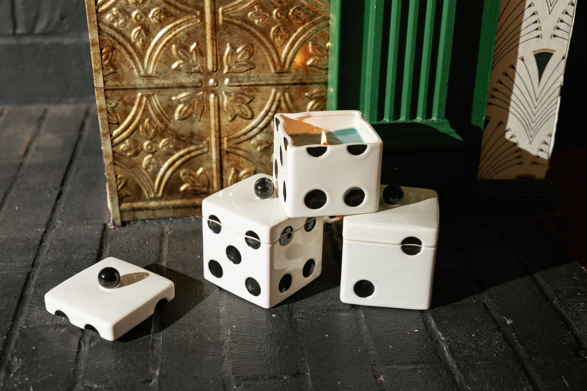 Three white, cube-shaped Misted Lime - Lucky Dice 10 oz Candle ceramic vessels with black dots, two of which have lids, with one candle burning inside the top container.