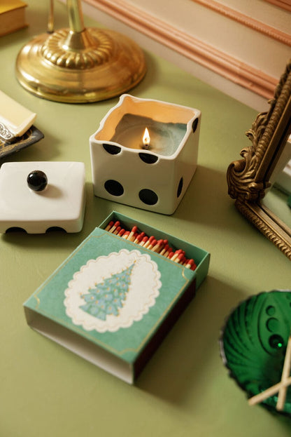 A lit candle in a dice-shaped holder sits beside Christmas Tree - Square Boxed Matches on a green surface, with a gold lamp, mirror, and green coaster—perfect for festive stocking stuffers.