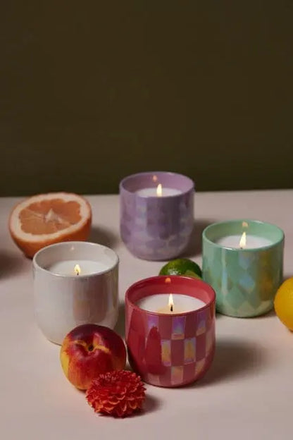 Four lit candles, including the Tobacco Vanilla - Lustre Candle 10 oz in a reusable ceramic vessel, are arranged on a table with assorted fruits and a flower against a plain background.