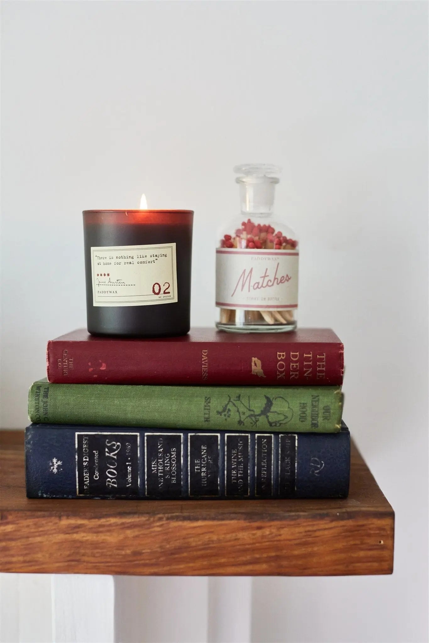 A Jane Austen - Library Candle 6 oz, a glass jar of matches, and three hardcover books sit on a wooden shelf against a plain white wall—a cozy nod to any Library Collection.