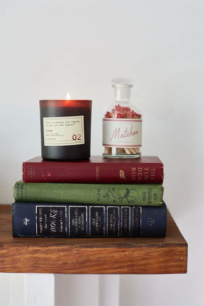 A Jane Austen - Library Candle 6 oz, a glass jar of matches, and three hardcover books sit on a wooden shelf against a plain white wall—a cozy nod to any Library Collection.