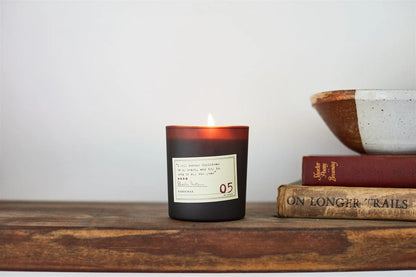 A lit Charles Dickens - Library Candle 6 oz sits on a wooden shelf next to a ceramic bowl and two stacked books, one red and one brown—adding a cozy touch to any Library Collection.