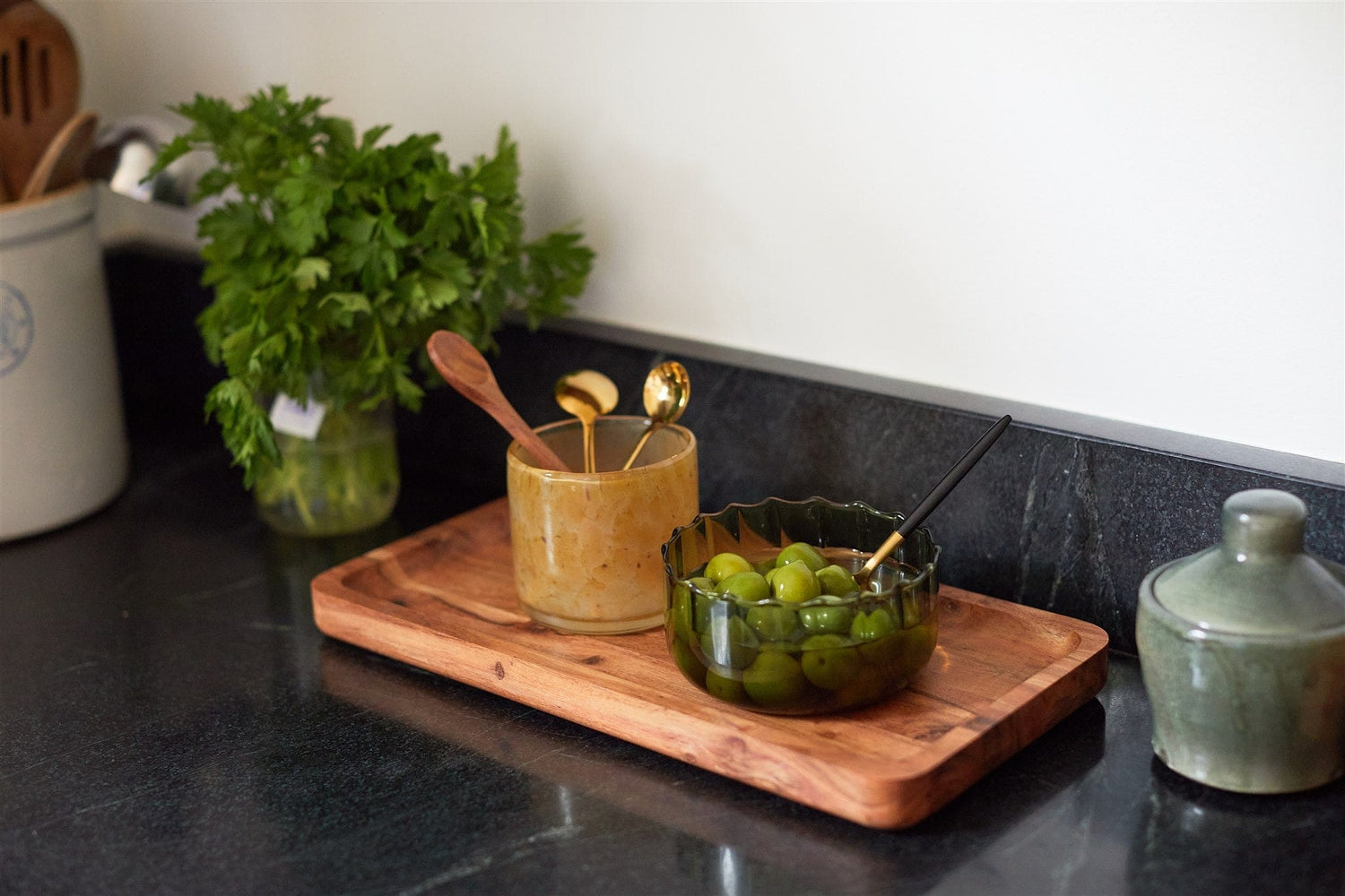 A wooden tray on a kitchen counter holds a glass jar with utensils and a bowl of green olives; fresh herbs are in a jar nearby.