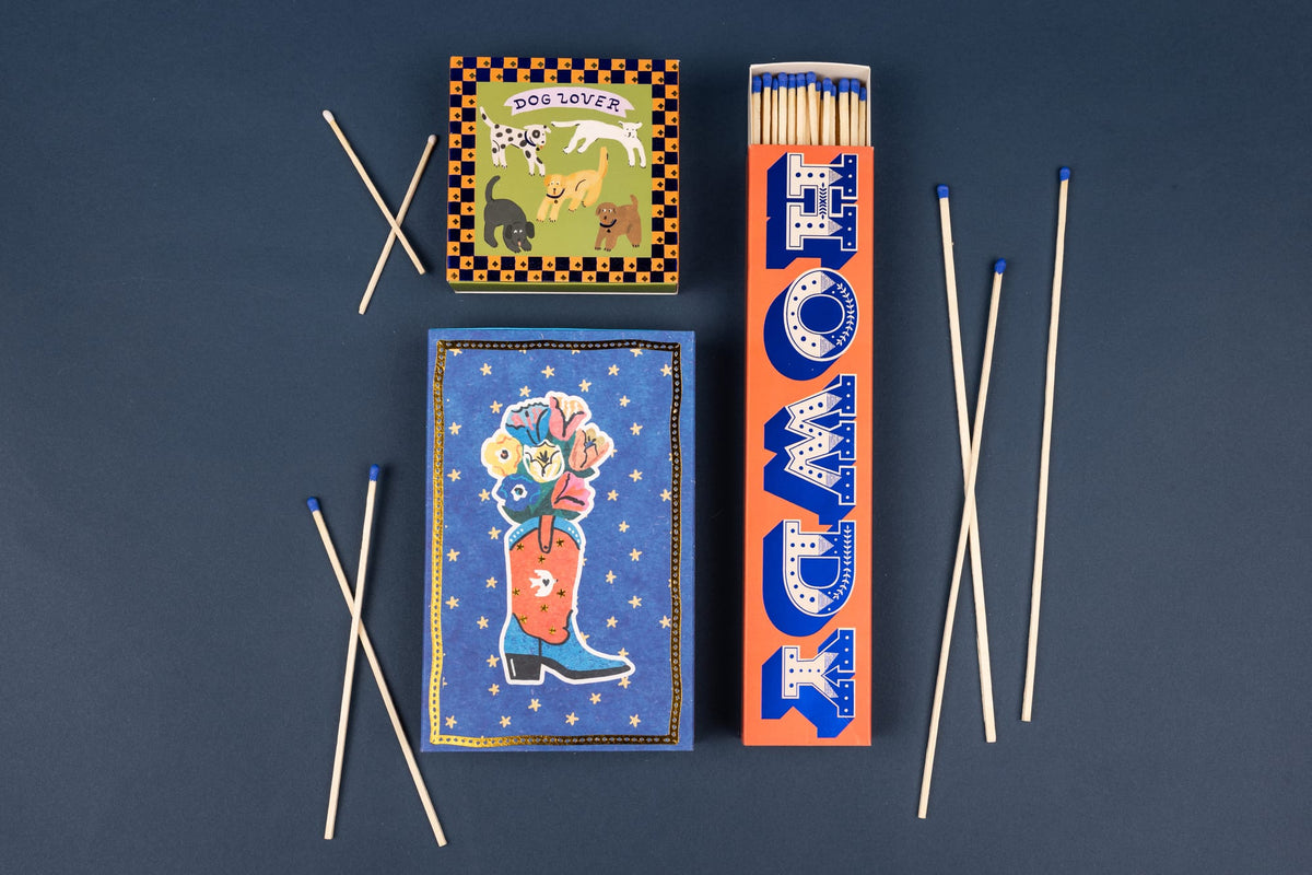 Three Howdy - Extra Long Boxed Matches decorated with illustrations of dogs, a flower-filled cowboy boot, and the word HOWDY, surrounded by several extra-long safety matches with blue tips.