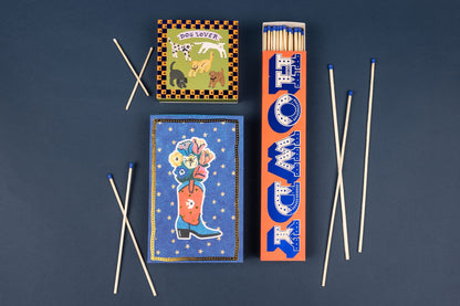 Three Howdy - Extra Long Boxed Matches decorated with illustrations of dogs, a flower-filled cowboy boot, and the word HOWDY, surrounded by several extra-long safety matches with blue tips.