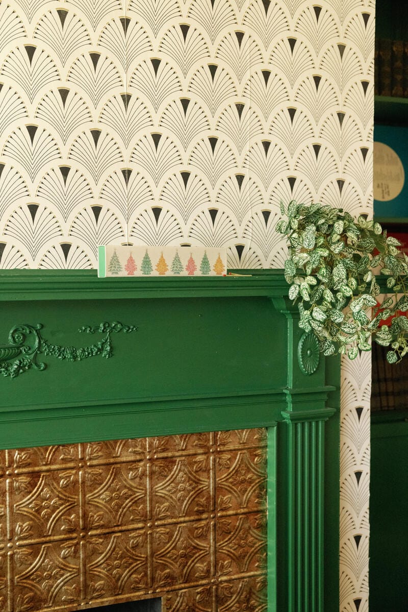 A green fireplace with a metallic tile inset, a leafy potted plant, and Christmas Trees - Extra Long Boxed Matches accented by a card with colorful trees sits against black and white art deco fan wallpaper.