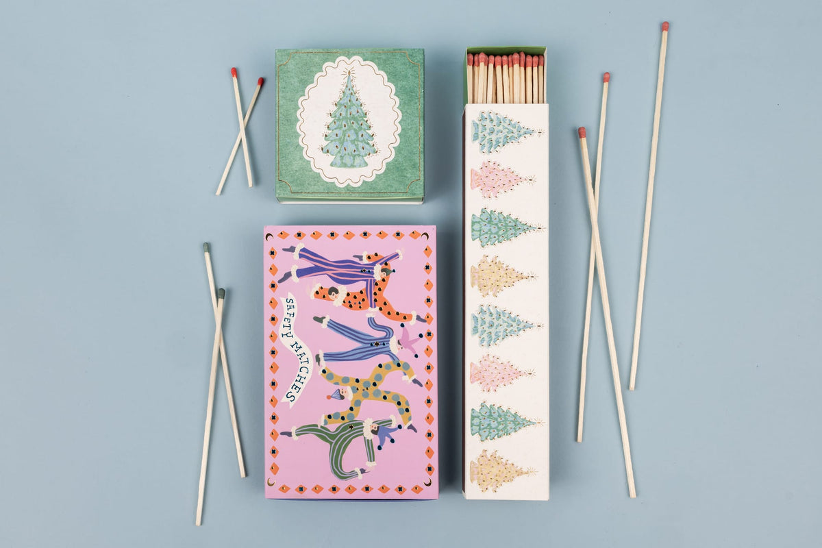Three Christmas Trees - Extra Long Boxed Matches boxes featuring illustrations of Christmas trees and figures in colorful patterned jumpsuits, accompanied by several loose extra-long safety matches.
