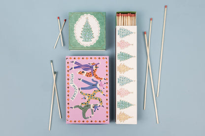 Three Christmas Trees - Extra Long Boxed Matches boxes featuring illustrations of Christmas trees and figures in colorful patterned jumpsuits, accompanied by several loose extra-long safety matches.