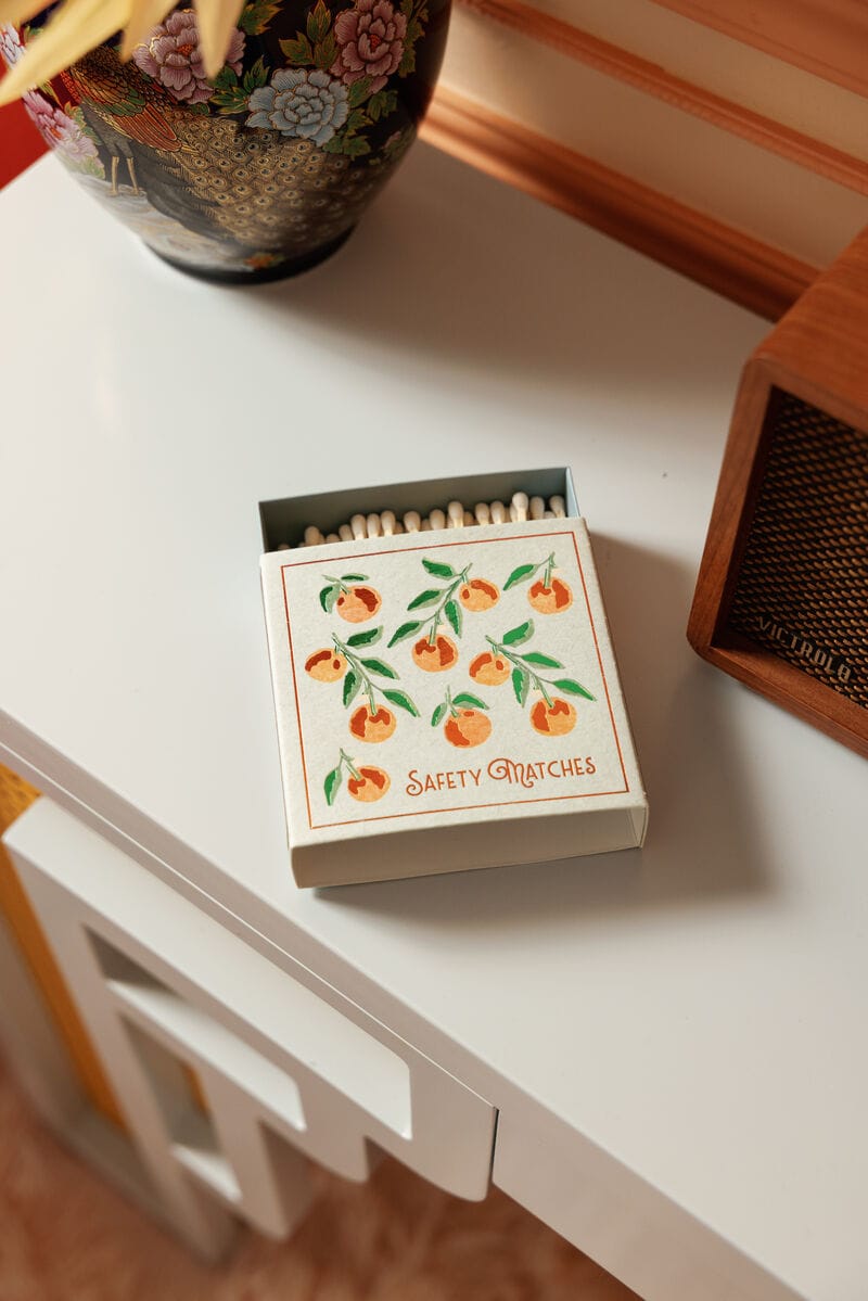 Oranges - Square Boxed Matches, featuring a hand-painted orange branch design, sits open on a white table next to a decorative vase and a wooden speaker.