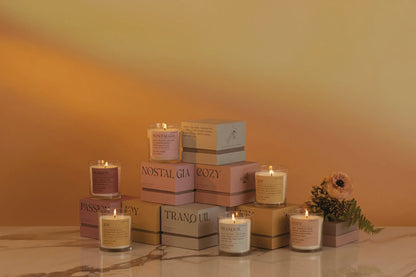 The Mood Collection 8oz Cashmere + French Orris "Cozy" candle sits in a glass jar on a pastel “Cozy” box, resting atop marble with a warm yellow gradient and floral accents.