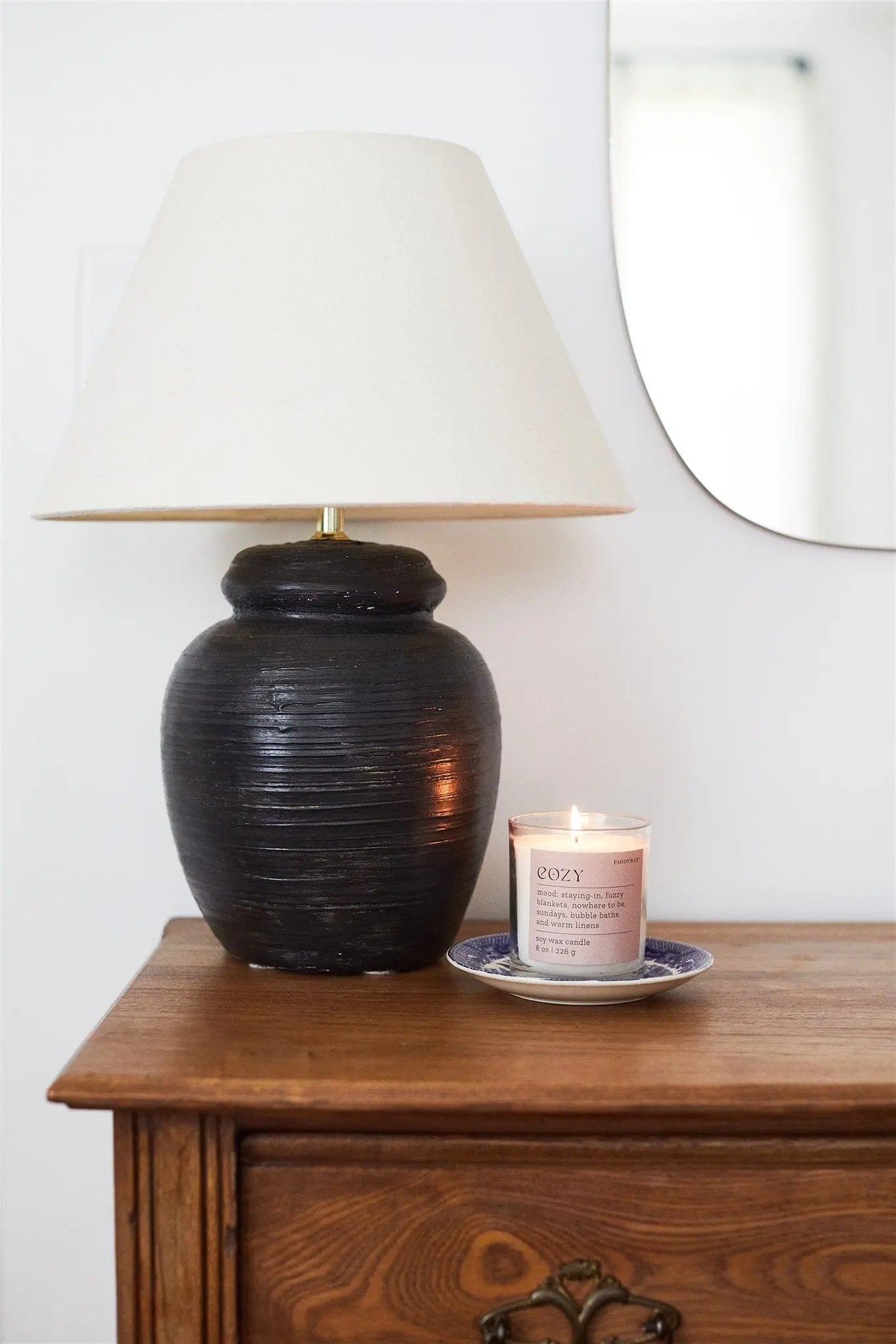A black ceramic table lamp with a white shade sits on a wooden dresser next to the Mood Collection 8oz - Cashmere + French Orris "Cozy" candle in a glass jar, resting on a blue and white saucer. A mirror hangs in the background.