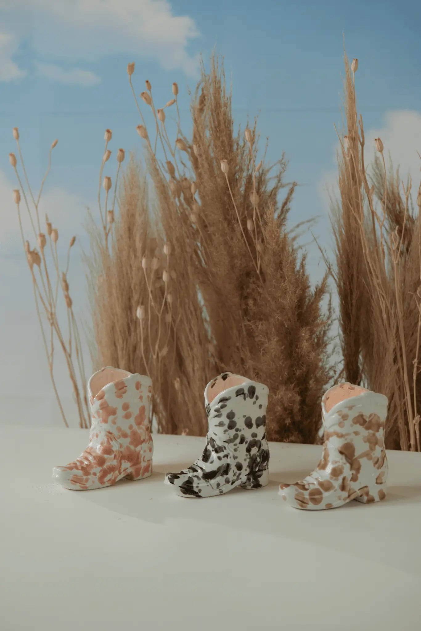Three small Nashville Boot 6 oz candles in Palo Santo Suede scent, with pink, black, and brown cow-print patterns, are displayed on a white surface with tall dried grasses and a blue sky backdrop for a Western-inspired vibe.