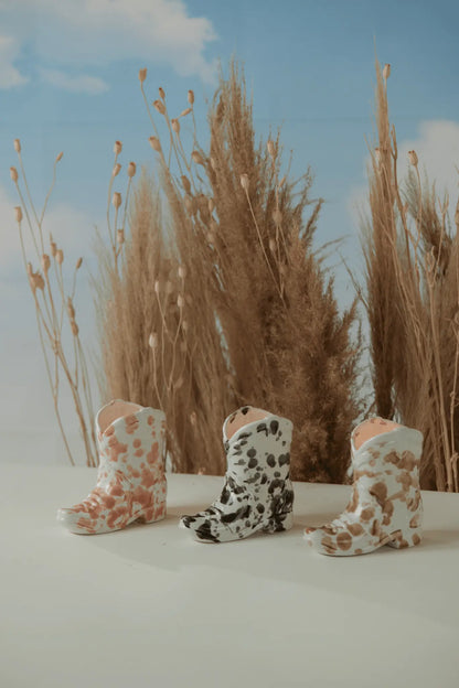 Three small Nashville Boot 6 oz candles in Palo Santo Suede scent, with pink, black, and brown cow-print patterns, are displayed on a white surface with tall dried grasses and a blue sky backdrop for a Western-inspired vibe.