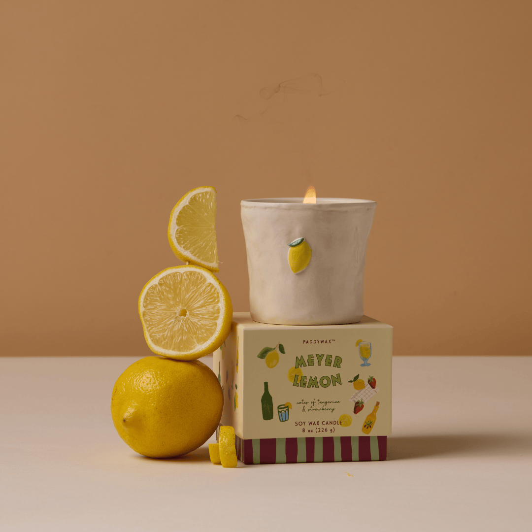 A lit Meyer Lemon - Bistro 8 oz Candle with a lemon design sits on its box beside a whole lemon and slices, set against a beige background.