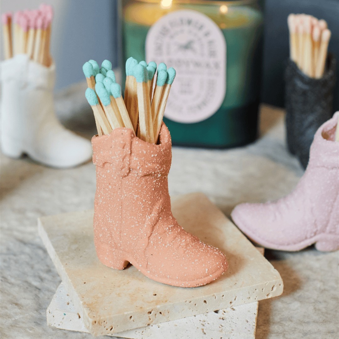 The Cowboy Boot Match Holder - Terracotta with blue-tipped matches sits on stone coasters, a candle and other boot holders in the background—an ideal Nashville gift or fun Western home decor accent.