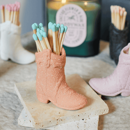 The Cowboy Boot Match Holder - Terracotta with blue-tipped matches sits on stone coasters, a candle and other boot holders in the background—an ideal Nashville gift or fun Western home decor accent.