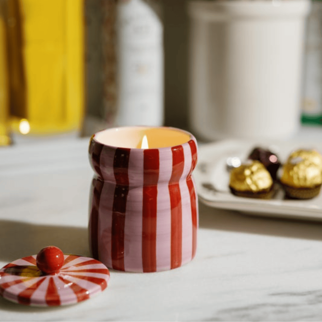 A Pink Peppermint - Cabana 6.5 oz Candle burns on the countertop beside assorted chocolates, its lid nearby. The hand-painted striped vessel adds charm to the cozy scene.