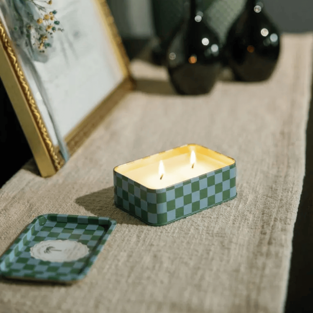 The Lush Palms - A Dopo 6.5 oz rectangular checkered tin candle with two lit wicks sits on a beige cloth, filling the air with a Mediterranean scent; its lid rests beside it on the table.