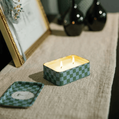 The Lush Palms - A Dopo 6.5 oz rectangular checkered tin candle with two lit wicks sits on a beige cloth, filling the air with a Mediterranean scent; its lid rests beside it on the table.