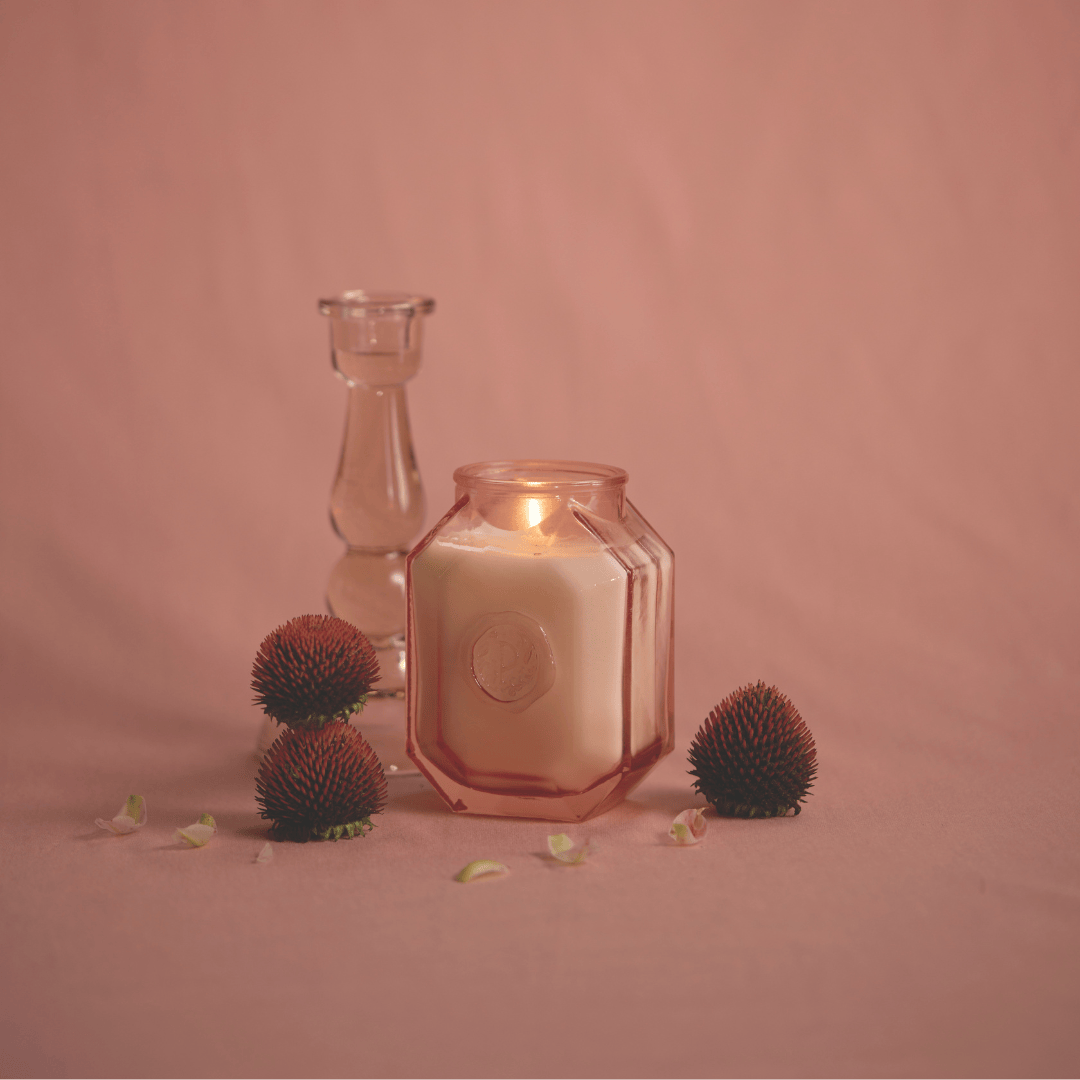 The Saffron Rose - Botanica 8 oz Candle glows in a hexagonal glass jar, displayed with a clear candlestick, dried flowers, and petals on a pink surface and background.