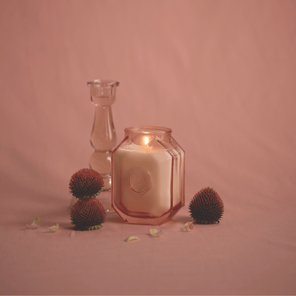 The Saffron Rose - Botanica 8 oz Candle glows in a hexagonal glass jar, displayed with a clear candlestick, dried flowers, and petals on a pink surface and background.