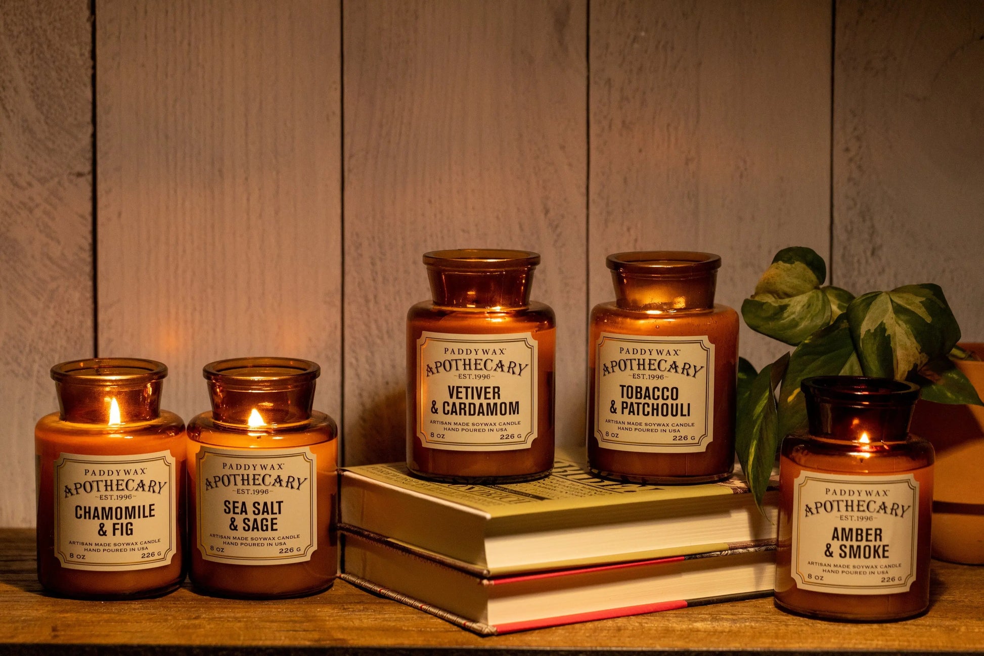 Three lit and two unlit Tobacco + Patchouli - Apothecary 8 oz candles in amber glass are arranged on stacked books and beside a plant, set against a wooden background.