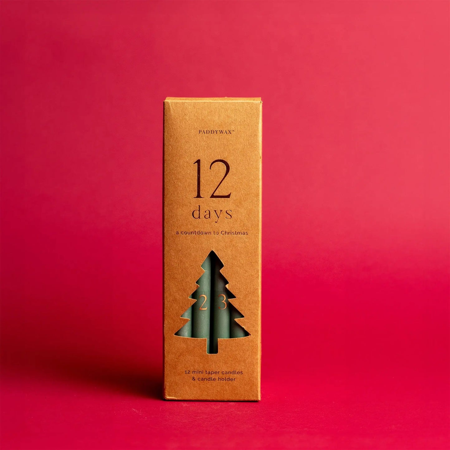 A brown box featuring the Cypress + Fir Taper 12 Days of Christmas Countdown Candle Set displays a cut-out tree shape revealing green taper candles inside, set against a solid red background—perfect for holiday gifting.