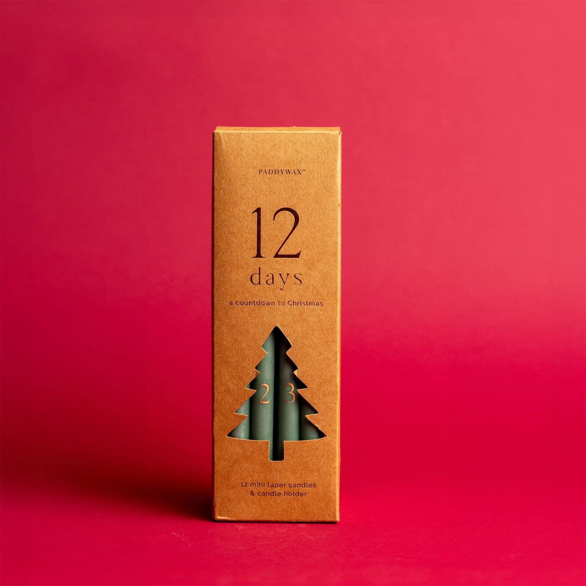 A brown box featuring the Cypress + Fir Taper 12 Days of Christmas Countdown Candle Set displays a cut-out tree shape revealing green taper candles inside, set against a solid red background—perfect for holiday gifting.