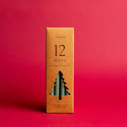 A brown box featuring the Cypress + Fir Taper 12 Days of Christmas Countdown Candle Set displays a cut-out tree shape revealing green taper candles inside, set against a solid red background—perfect for holiday gifting.