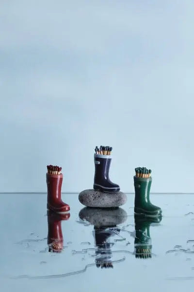 A navy blue ceramic Wellington Boot match holder, filled with safety matches, is displayed on a stone atop a reflective surface with a light blue background.