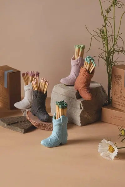 The Cowboy Boot Match Holder in light blue is filled with matches and displayed on stone blocks, surrounded by its packaging and a white flower for a vintage-inspired look against a neutral background.
