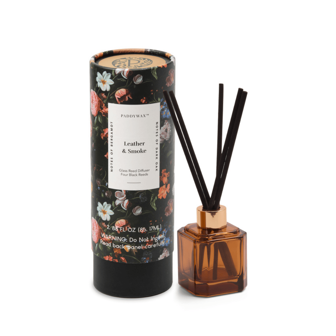 The Leather + Smoke - Botanica Diffuser 2.88 oz features black reeds in a brown glass bottle and floral-patterned packaging—a refined, modern addition to your home fragrance collection.