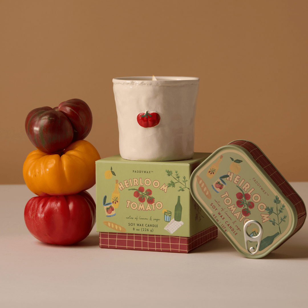 The Heirloom Tomato - Bistro 8 oz Candle, featuring a tomato design, sits on a decorative "Heirloom Tomato" box next to a stack of three tomatoes and a similarly branded tin.