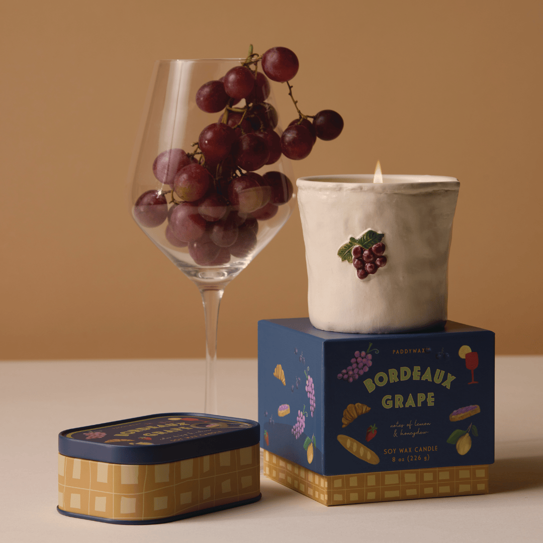 A glass of red grapes, the Bordeaux Grape - Bistro 8 oz Candle with a grape design, and two blue candle boxes are displayed on a beige surface against a brown background.