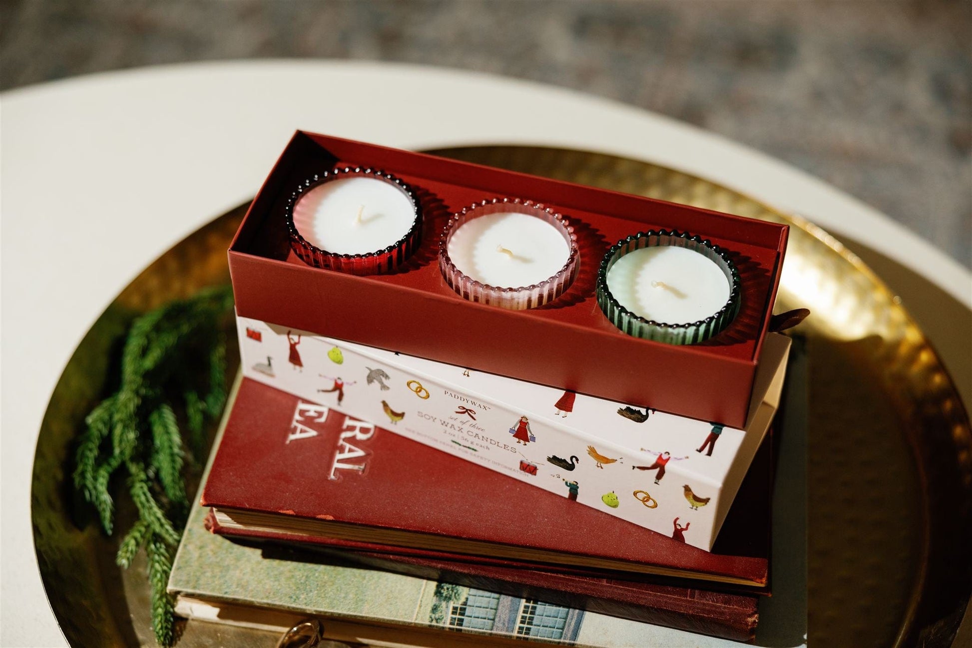 The 12 Days Of Christmas Holiday Set - 2 oz features three festive scented candles in colorful holders, displayed on a stack of books atop a round gold tray, with green decorative foliage nearby.