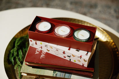 The 12 Days Of Christmas Holiday Set - 2 oz features three festive scented candles in colorful holders, displayed on a stack of books atop a round gold tray, with green decorative foliage nearby.