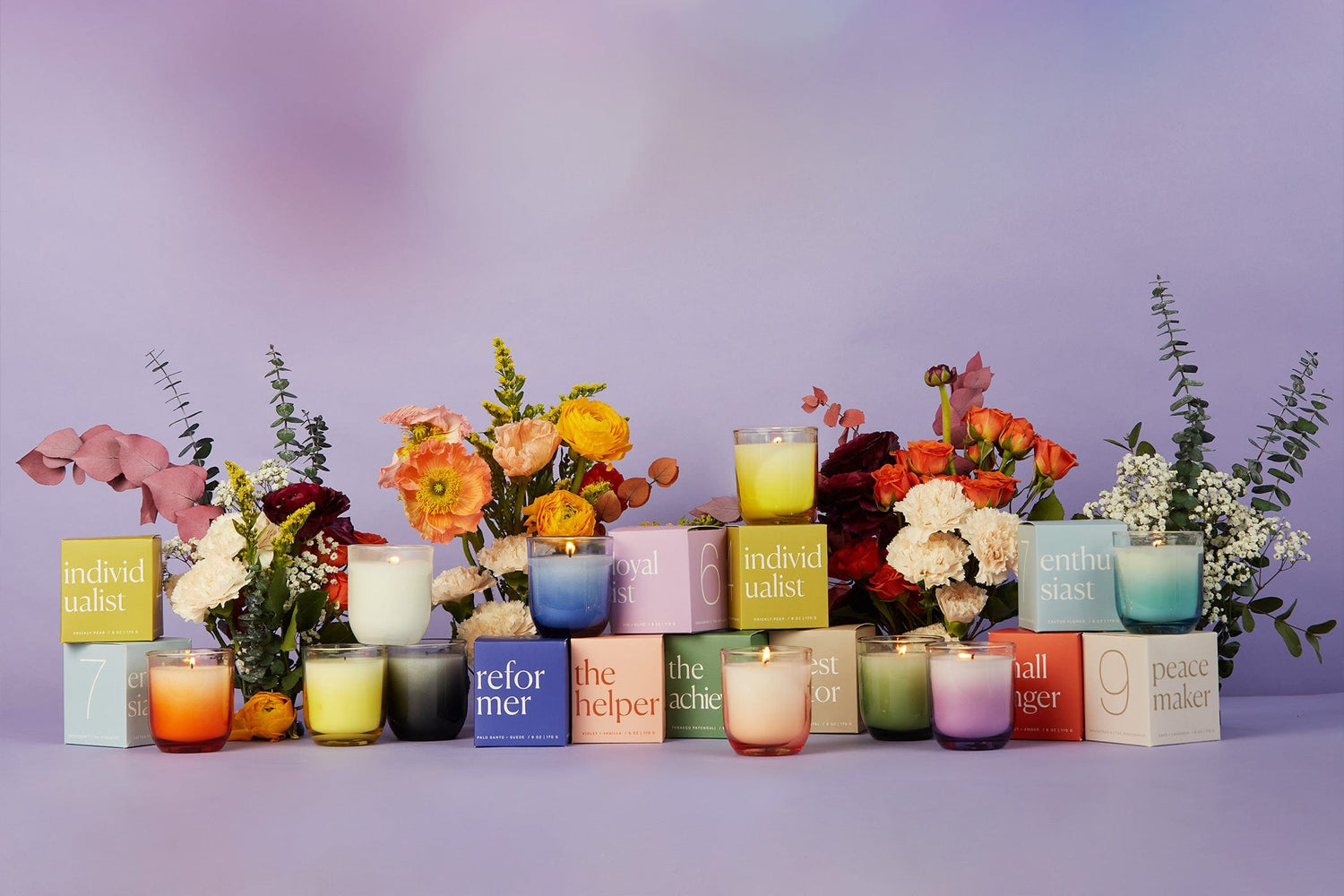 A colorful display of candles in glass jars, each labeled with different personality types, surrounded by vibrant floral arrangements against a soft purple background.